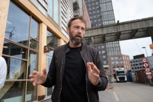 Architect Anders Lendager with a graying beard and dark bomber jacket gesturing with both hands as he speaks in front of his timber-clad TRÆ office building in the Sydhavnen port district of Aarhus, Denmark, with the wooden facade and large windows visible on the left and modern high-rise buildings, a construction crane, and a freight truck in the background under an overcast sky
